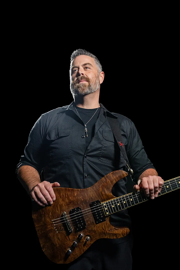 Jake O'Donnell holding a wood-finished guitar against a dark stage background